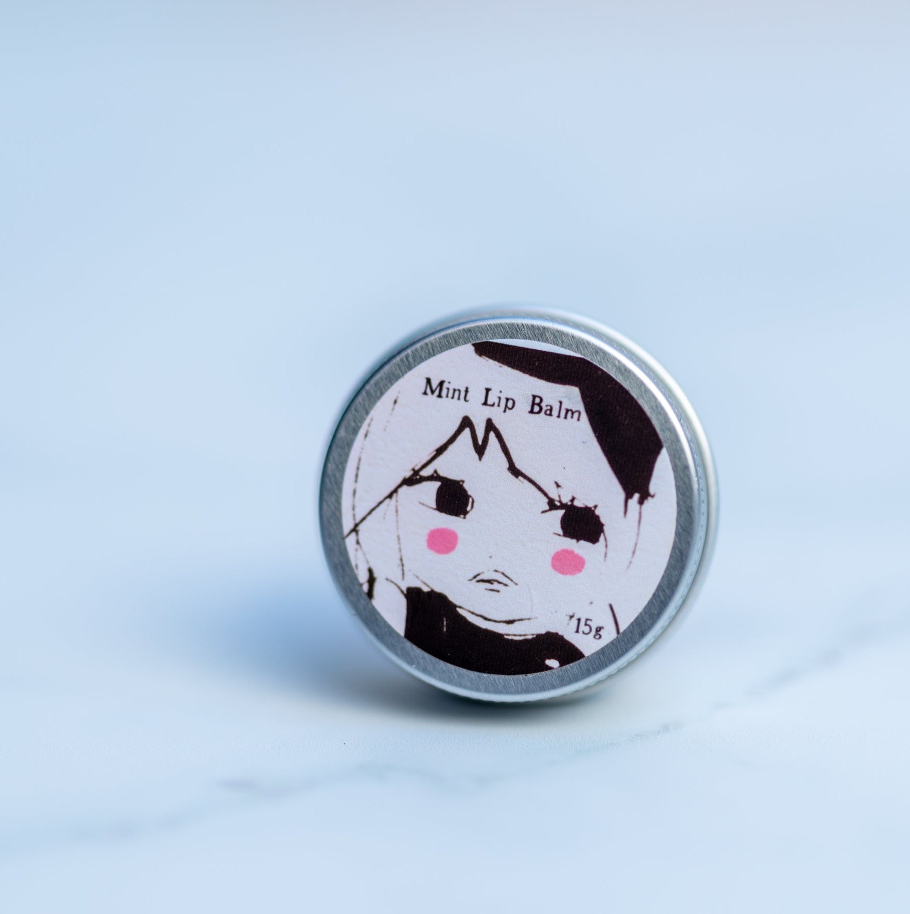 Aluminium tin of lip balm with a screen printed label of a girl with a big hair bow and pink rosey cheeks on a marble background
