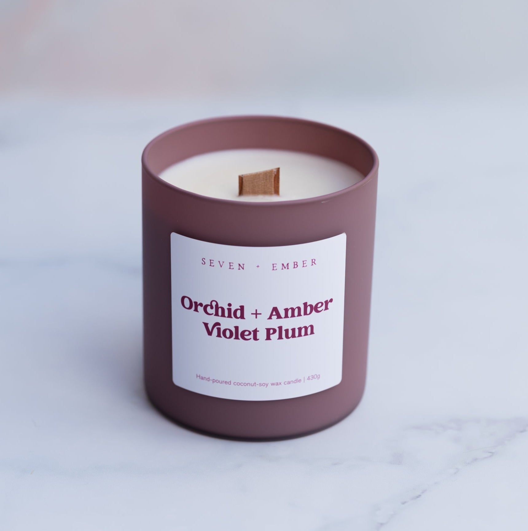 Orchid and amber canlde with wooden wick in a plum coloured frosted glass vessel on a blurred marble background