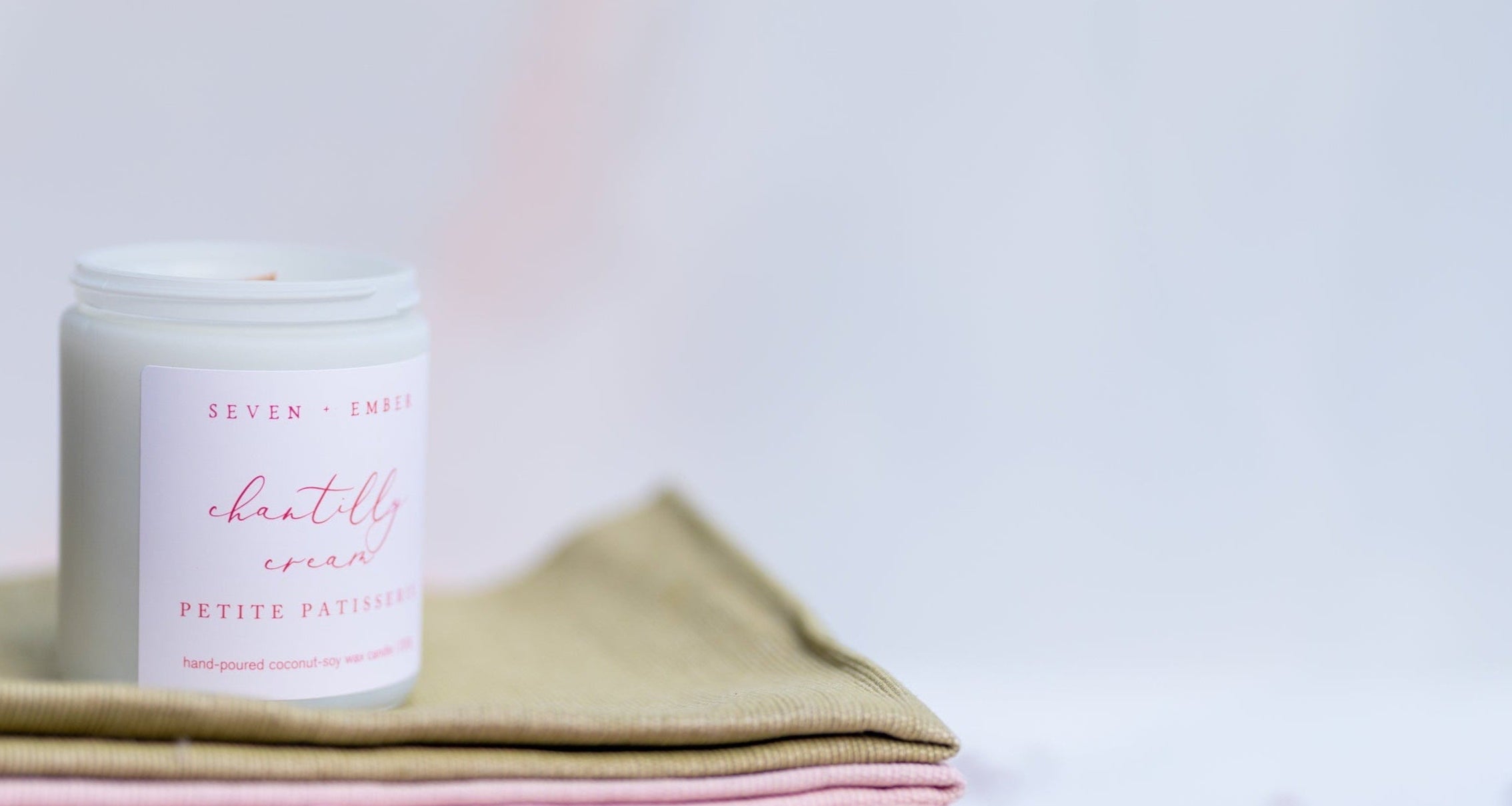 Chantilly cream candle in a white vessel on a green and pink cloth on a white blurred background
