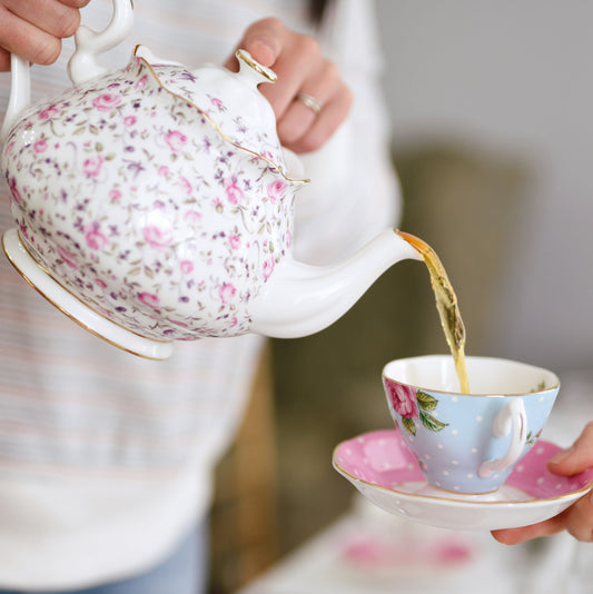 A woman pouring a cup of tea from a floral adorned teapot into a pale blue floral teacup