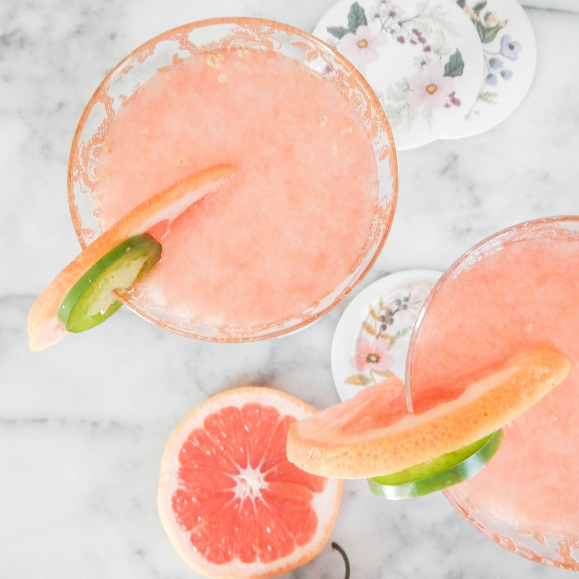 Refreshing grapefruit drinks in glasses garnished with grapefruit and lime slices on a marble background with floral coasters