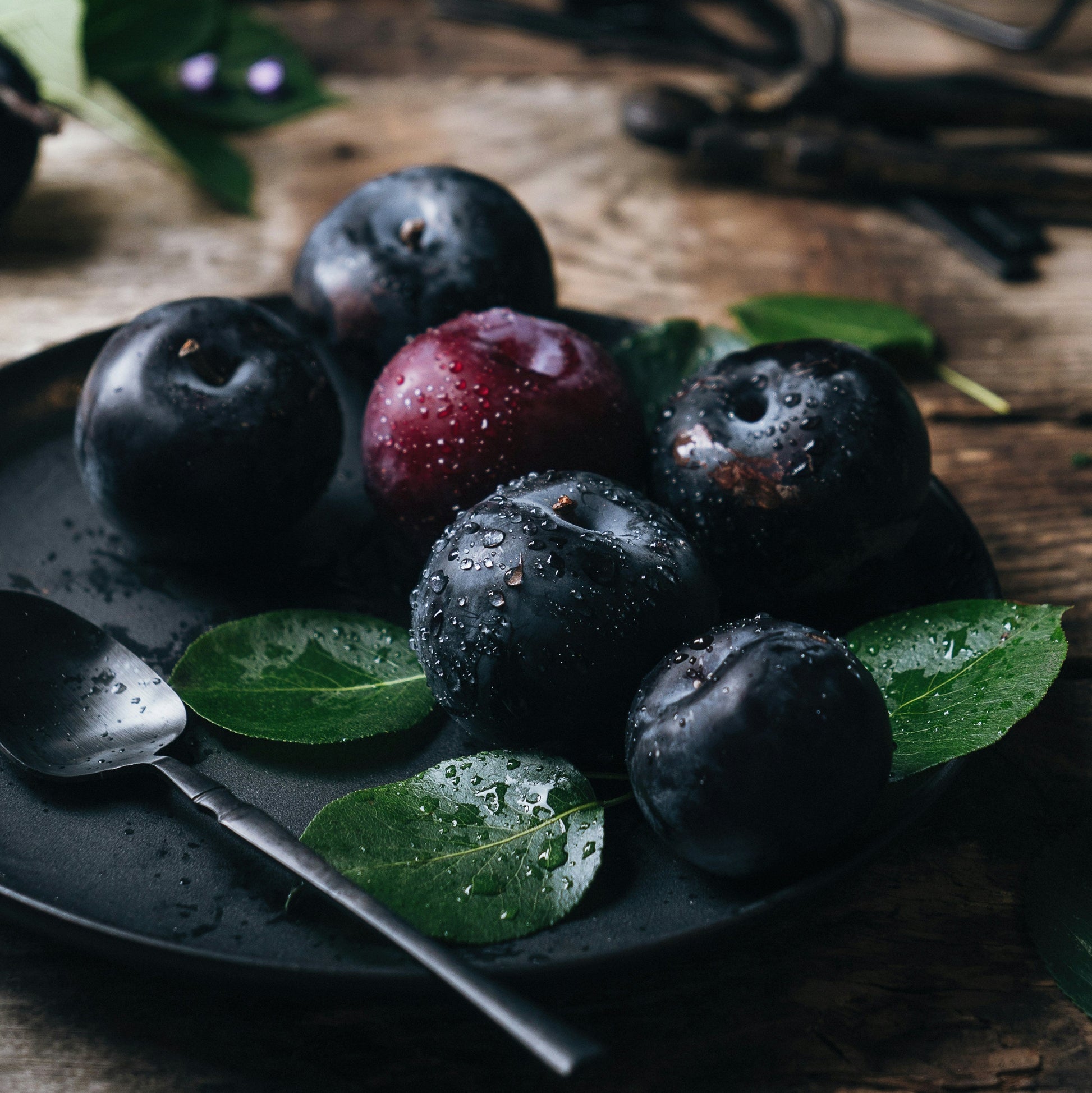 A black plate with blood plums, green leaves and aspoon with vanilla beans in the background on a wooden surface