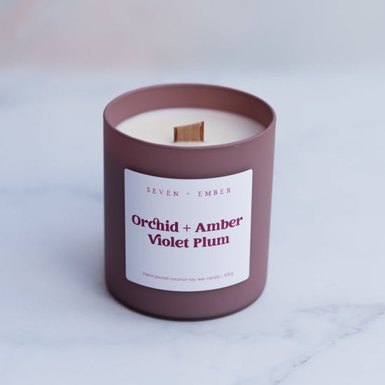 Orchid and amber canlde with wooden wick in a plum coloured frosted glass vessel on a blurred marble background