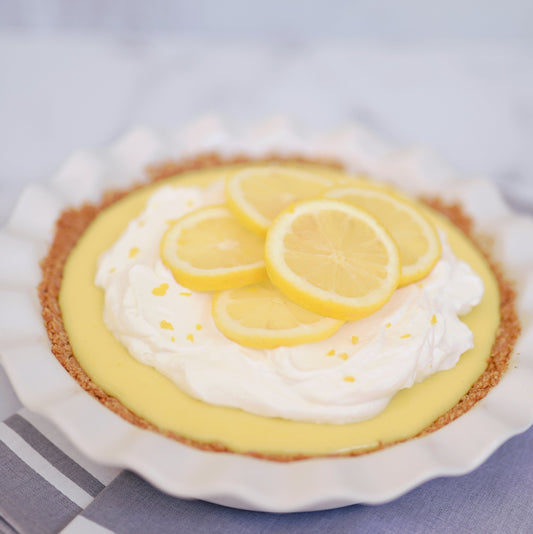Lemon Meringue tart in a white fluted pie dish topped with cream and slices of lemon on a white background with blue tea towels in the foreground