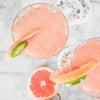 Refreshing grapefruit drinks in glasses garnished with grapefruit and lime slices on a marble background with floral coasters