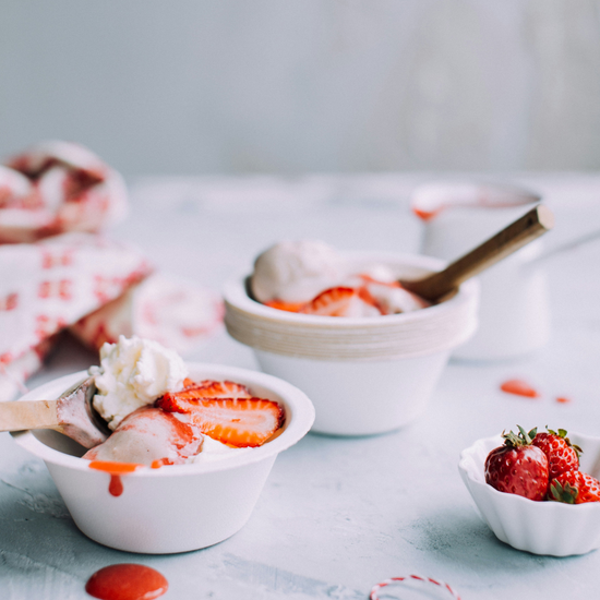 Mini bowls of strawberries and cream with mini wooden spoons and a checkered cloth and a mini bowl of red strawberries