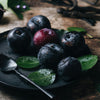 A black plate with blood plums, green leaves and aspoon with vanilla beans in the background on a wooden surface