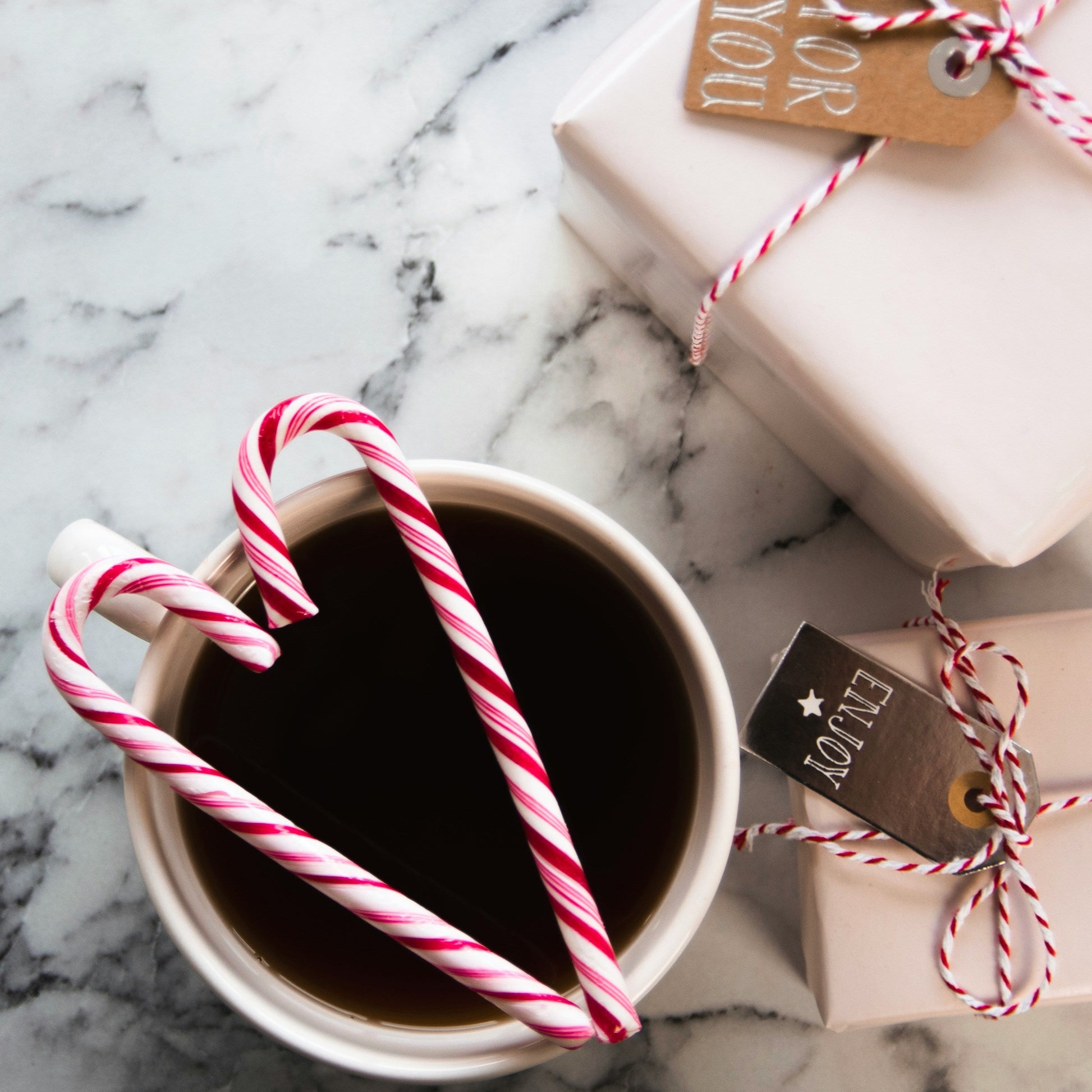 A festive image of two candy canes in the shape of a love heart atop a cup of hot chocolate on a merble surface with two Christmas gifts wrapped in red and white twine with a gold tag
