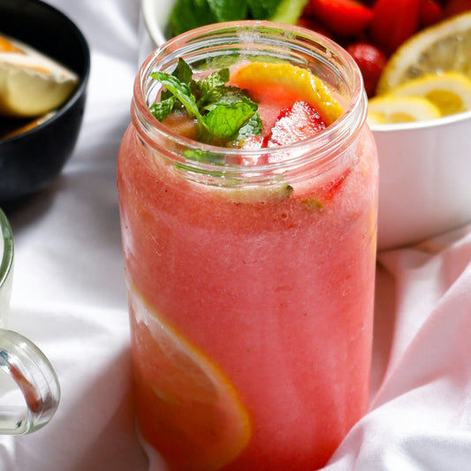 A refreshing glass of watermelon juice with orange slices and a sprig of mint and a bowl of lemon slices and cherries in the background
